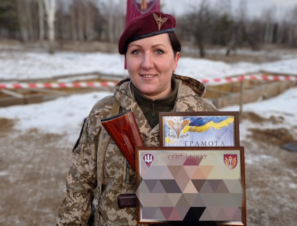 Військовослужбовиця у береті ДШВ тримає грамоту та відзнаки, отримані за службу чи досягнення. Урочистий момент підкреслює визнання її професійних заслуг і внеску у військову справу.