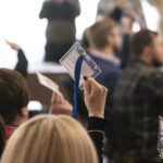 Учасники заходу піднімають картки для голосування, беручи участь у процедурі ухвалення рішень. Фото передає момент активного голосування в залі під час офіційного зібрання.