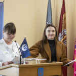 Жінки біля трибуни беруть участь у церемонії оголошення результатів або вручення документів. Атмосфера події залишається урочистою та організованою.
