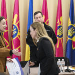 Молода представниця організації вручає документи або сертифікат учасниці заходу. На фоні державні та військові прапори підсилюють офіційність церемонії.