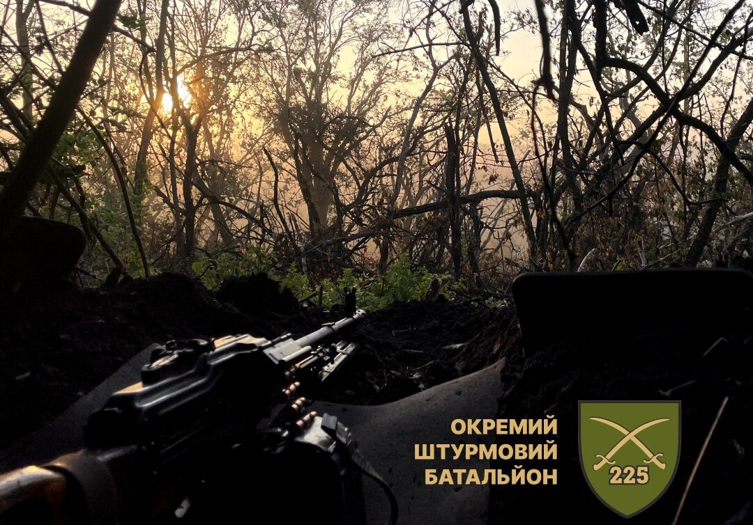 На фото — бойова позиція серед густої лісосмуги на світанку, де крізь переплетіння дерев пробивається м’яке сонячне світло. На передньому плані видно кулемет, готовий до ведення вогню, а емблема 225-го окремого штурмового батальйону підкреслює належність світлини до бойової роботи підрозділу.