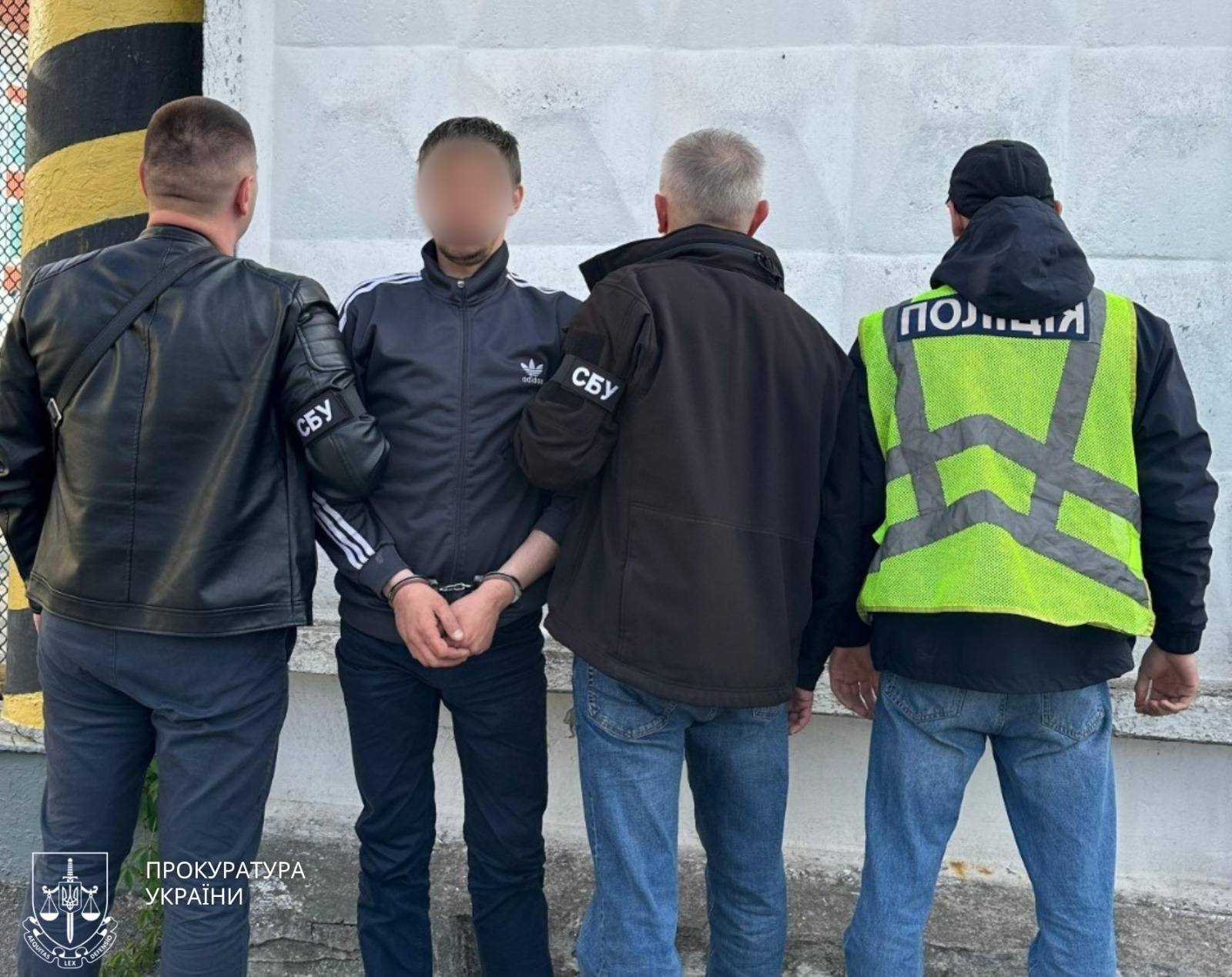 Затриманий стоїть в оточенні правоохоронців на тлі бетонного паркану