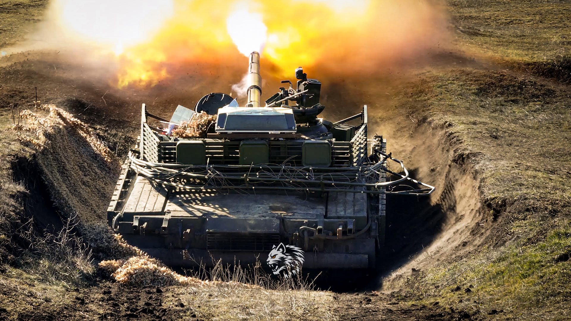 Танк веде інтенсивний вогонь із підготовленої вогневої позиції, здіймаючи потужний спалах, пил і полум’я навколо гармати. Замаскована техніка розташована в укріпленому окопі серед відкритої місцевості, що підкреслює бойовий характер дій.