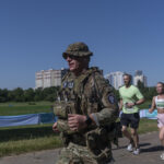 Військовий у повному спорядженні біжить дистанцію поруч із цивільними учасниками забігу. На задньому плані видно зелену зону та багатоповерхівки, що підкреслює міський формат заходу.
