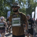 Учасник у тактичному спорядженні біжить у загальній колоні, на його грудях закріплений стартовий номер забігу імені «Рекрута». Поруч рухаються інші бігуни, а навколо видно прапори партнерів та організаторів.