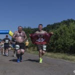 Двоє учасників вибігають з-під арки, один із них тримає прапор із військовою емблемою та написом. Позаду видно інших бігунів, які продовжують дистанцію сонячною парковою трасою.