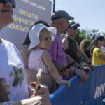 Глядачі стоять уздовж траси за огорожею та спостерігають за учасниками забігу. Серед них видно дитину в білому капелюшку, яка уважно дивиться на дистанцію.