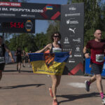 Учасниця забігу перетинає фінішну зону з прапором III окремої штурмової бригади. Поруч фінішують інші бігуни, а над трасою розміщена арка «Забіг честі ім. Рекрута 2026».