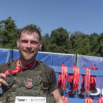 Усміхнений учасник у тактичному спорядженні тримає в руках медаль після забігу. За його спиною видно стенд із червоними стрічками нагород для фінішерів.