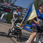 Учасниця з прапором проходить фінішну зону поруч з іншими бігунами. На задньому плані видно арку забігу, таймер і родину з дитячим візком.