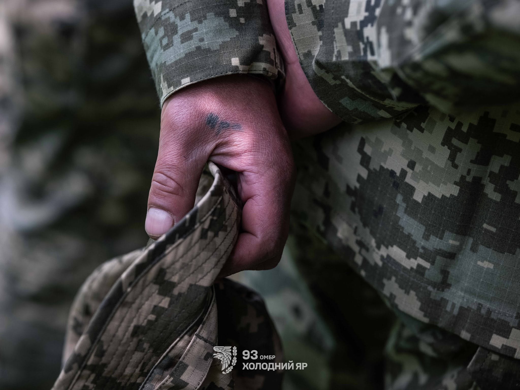 Крупним планом показана рука військового у камуфляжі, який стискає польовий кашкет. Зосередження на деталях — формі, руці й затемненому тлі — передає стриману напругу та буденну суворість військової служби.