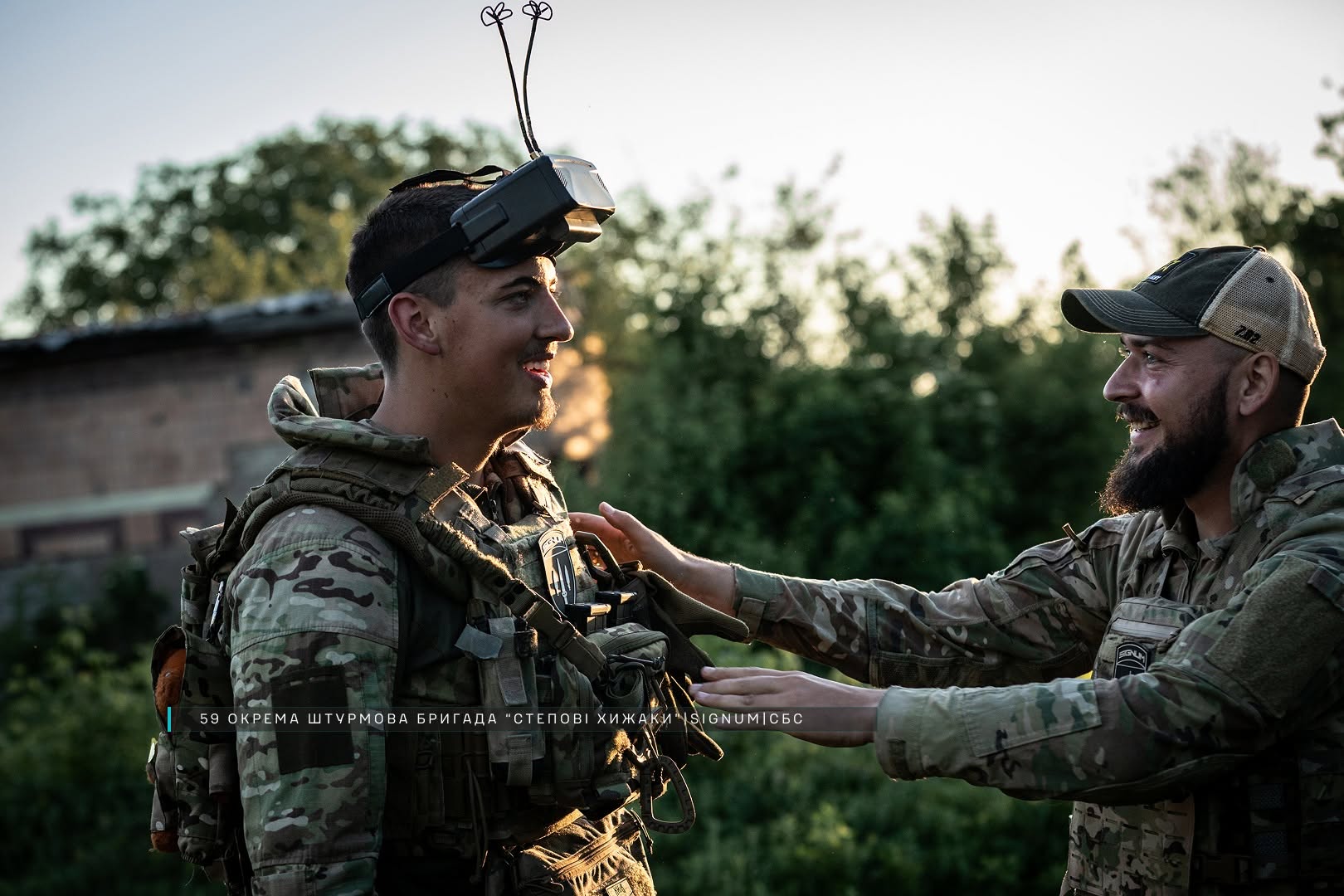 Двоє військових у камуфляжі усміхаються та спілкуються на тлі зелені й вечірнього світла. Один із них має на голові окуляри для керування FPV-дроном, а інший допомагає йому з екіпіруванням.