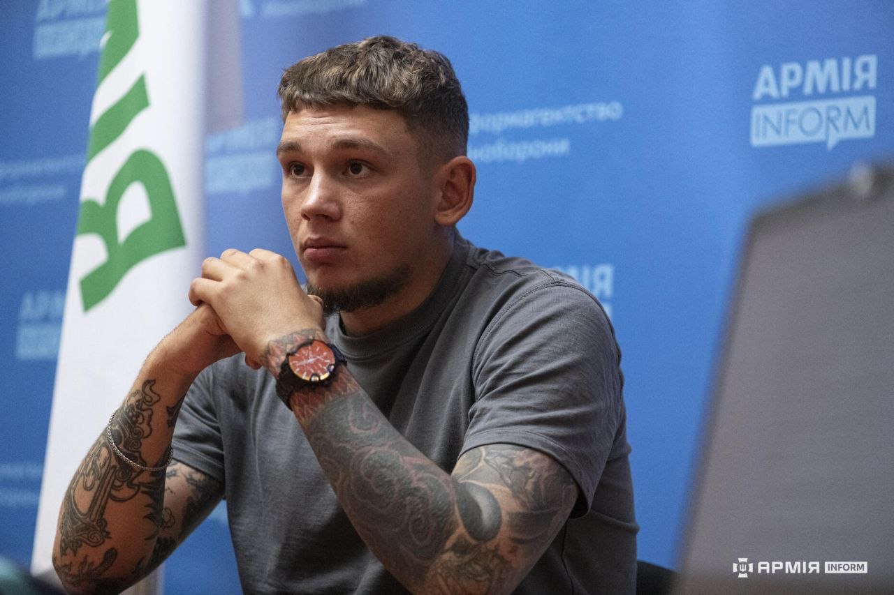 Військовослужбовець «Азову» Павло на позивний «Пінг» сидить за столом, склавши руки перед собою, та уважно слухає співрозмовника. Його руки вкриті татуюваннями, а позаду розміщені синій інформаційний банер і білий прапор із зеленим написом.