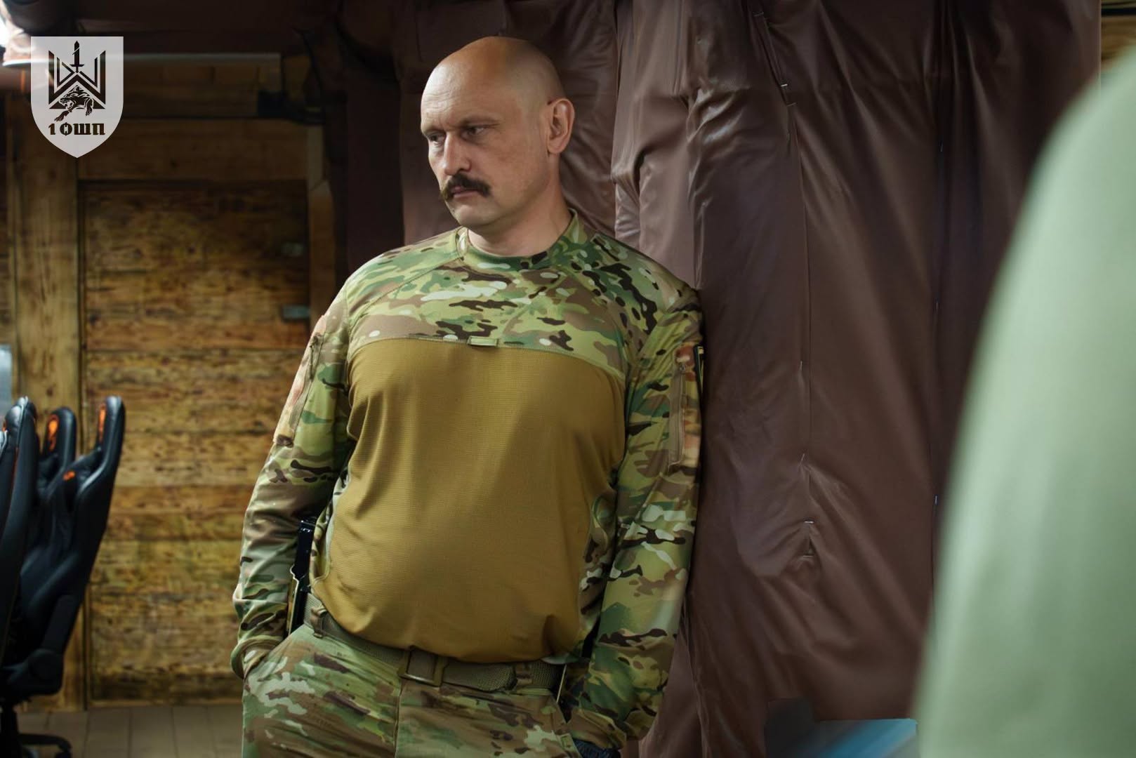 Військовий стоїть біля стіни у приміщенні, тримаючи руки в кишенях і зосереджено дивлячись убік. Навколо видно дерев’яні панелі, крісла та робочу атмосферу штабу або пункту управління.