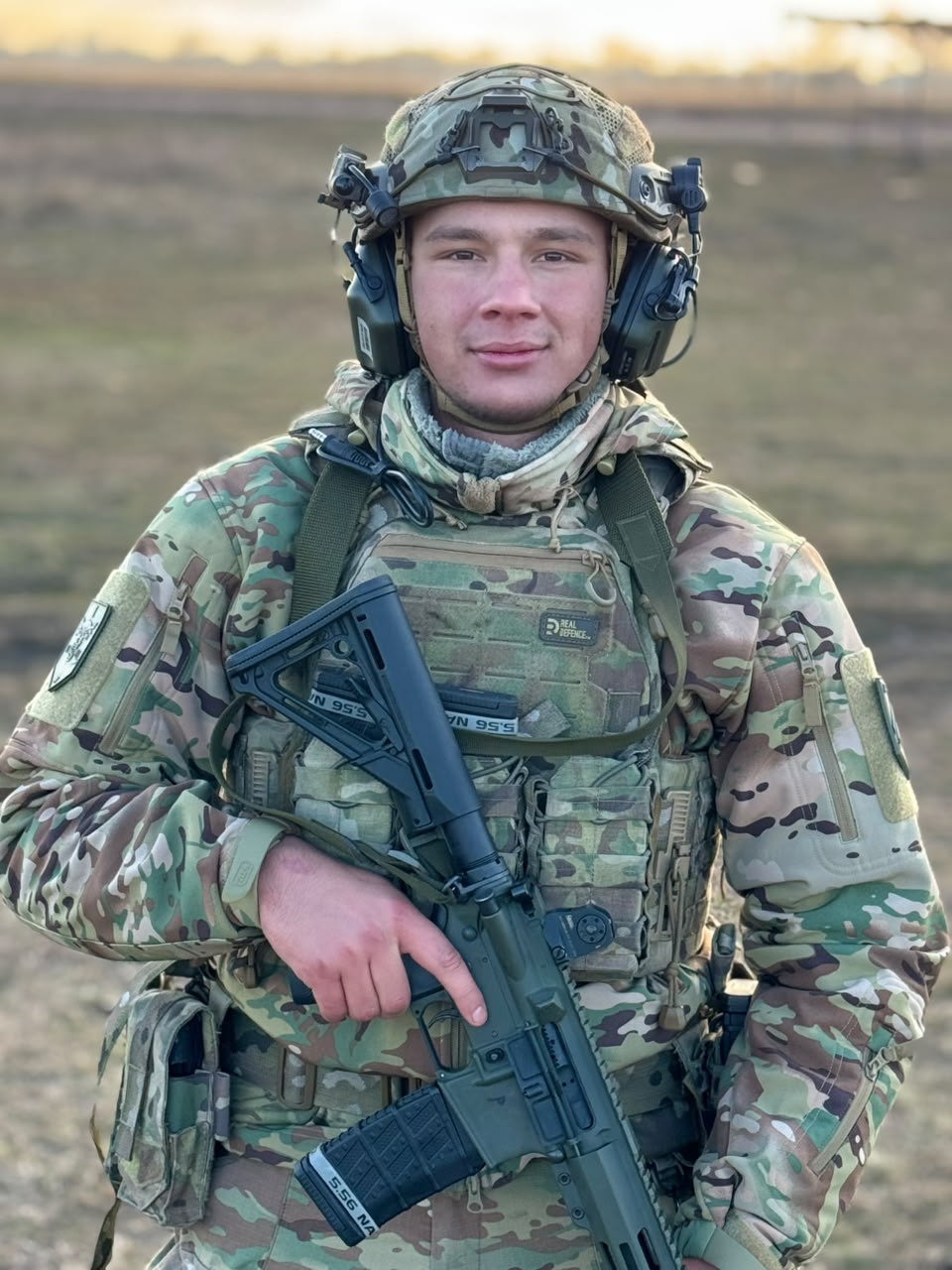 Військовослужбовець «Азову» Павло на позивний «Пінг» у камуфляжі, шоломі та захисному спорядженні стоїть на відкритій місцевості. Він тримає перед собою автоматичну гвинтівку та дивиться прямо перед собою.