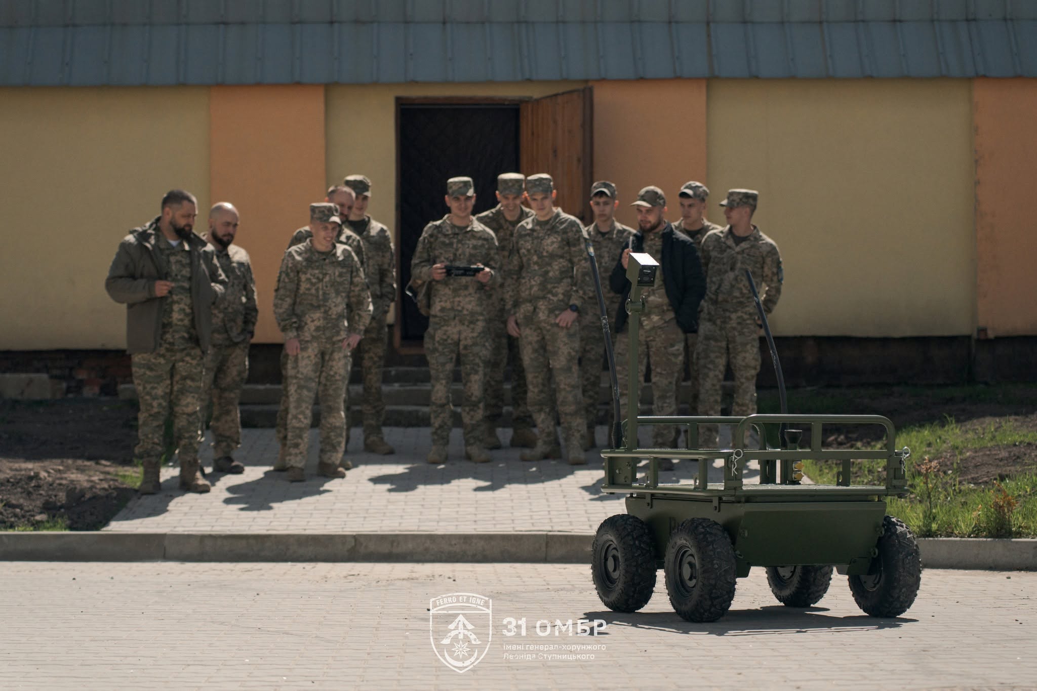 Група військовослужбовців спостерігає за роботою наземної роботизованої платформи на подвір’ї біля будівлі. Один із бійців керує технікою з пульта, демонструючи її можливості.