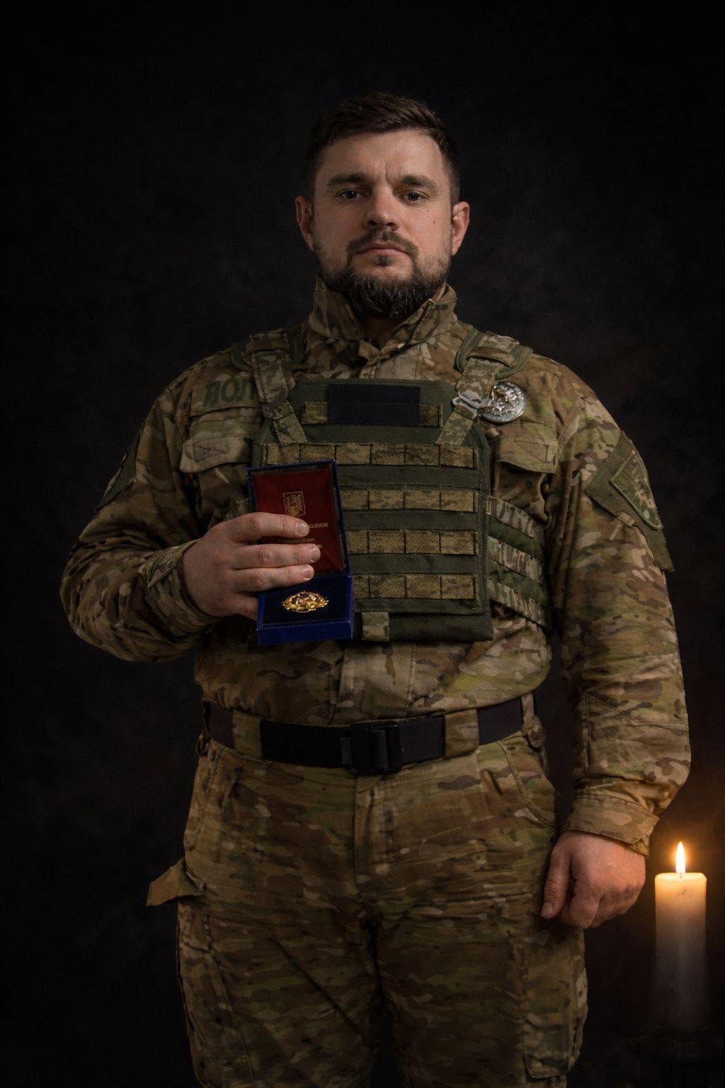 На портретному фото чоловік у камуфляжній формі тримає футляр із відзнакою. Темний фон і запалена свічка поруч створюють стриману, пам’ятну атмосферу.