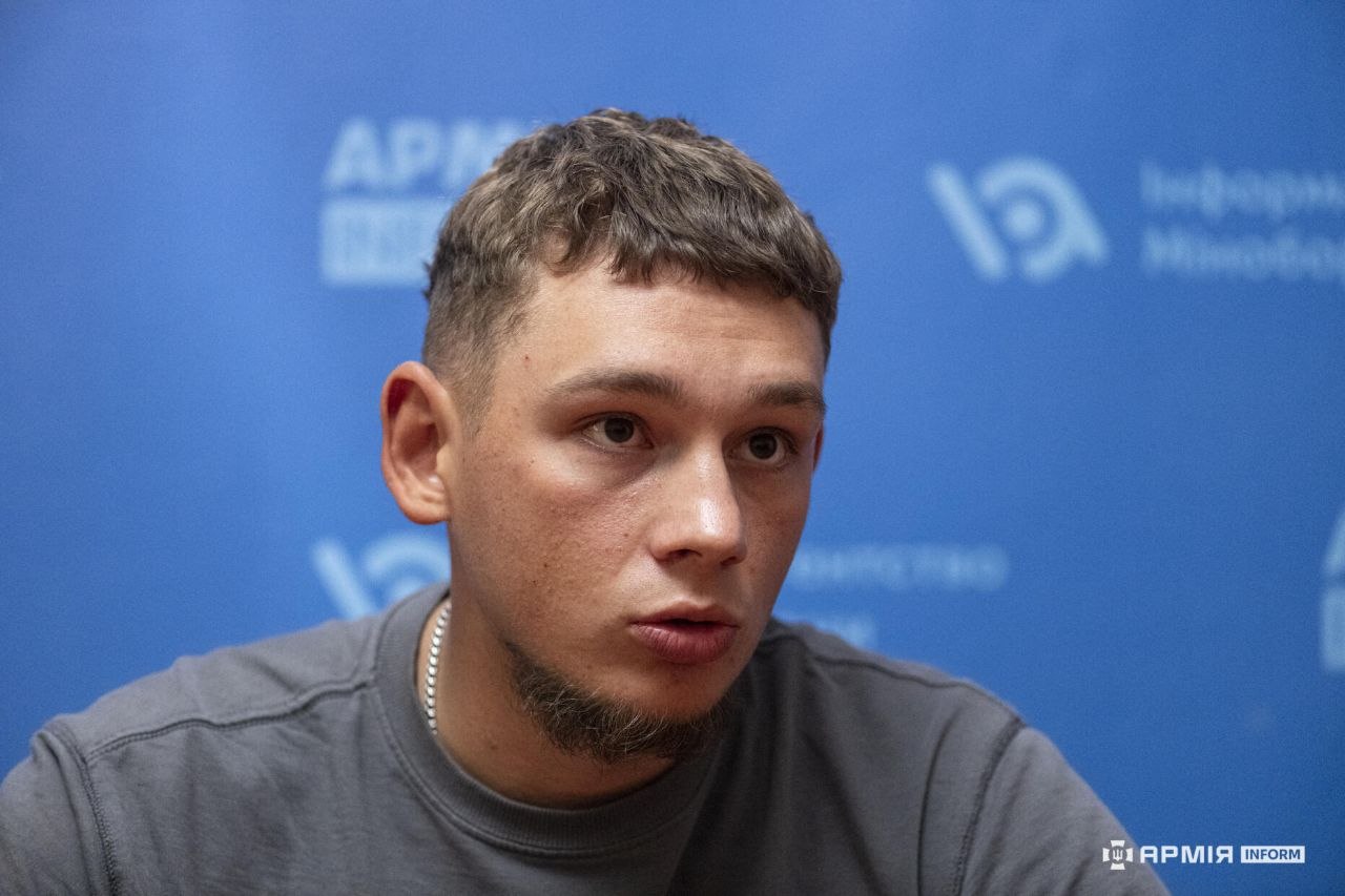 Чоловік під час розмови дивиться трохи вбік і промовляє відповідь. За його спиною розташований синій банер з емблемами інформаційного агентства.