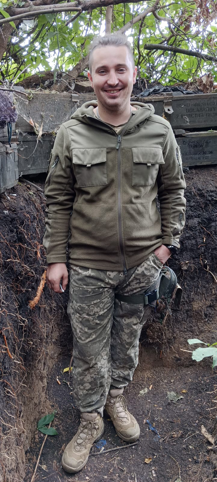 Військовий у польовій формі стоїть в укритті серед гілок і земляних стінок. Він усміхається, тримає руку в кишені, а поруч видно елементи спорядження.