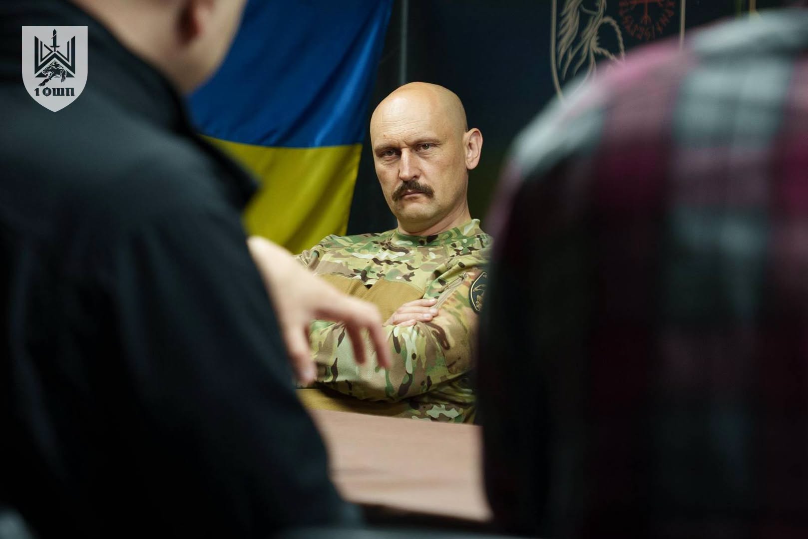 Військовий сидить за столом під час розмови, а на передньому плані видно силуети інших присутніх. Позаду нього розміщений український прапор, що підкреслює офіційний характер зустрічі.