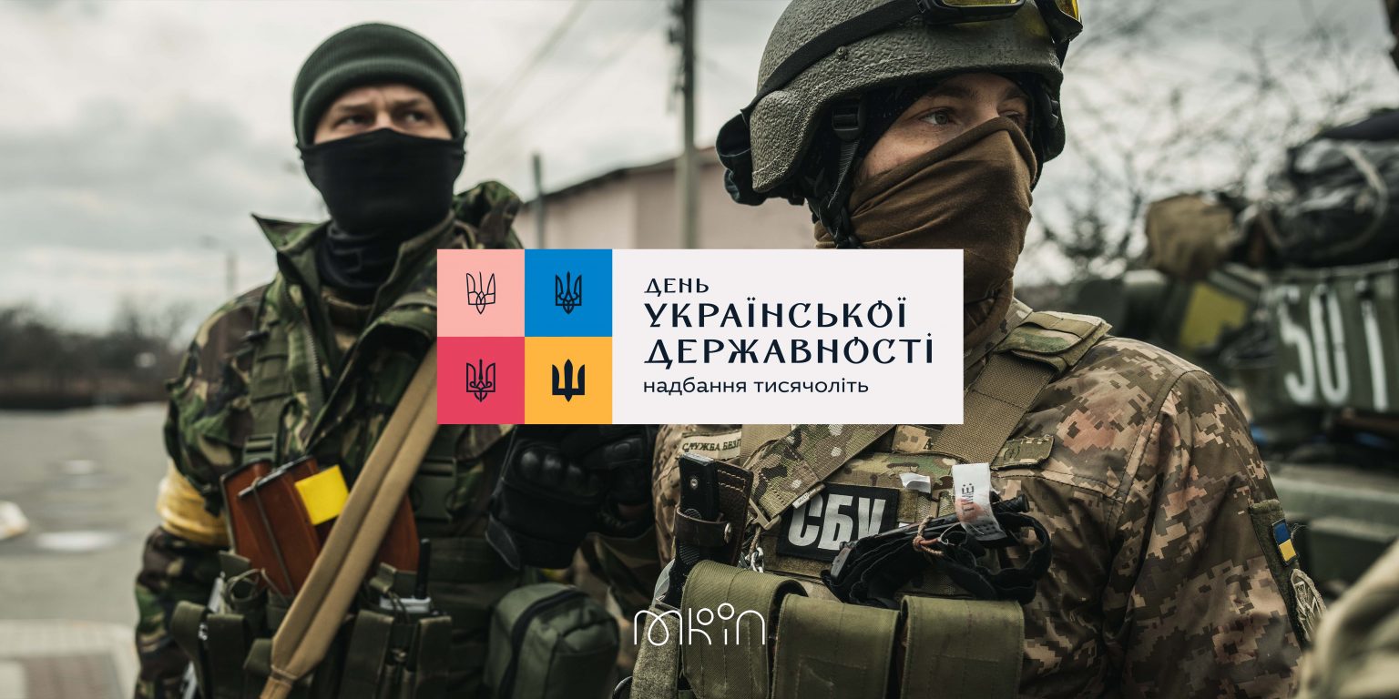 Двоє озброєних українських військовослужбовців у захисному спорядженні, поруч стоїть військова техніка. У центрі розміщено емблему Дня Української Державності з написом «Надбання тисячоліть».