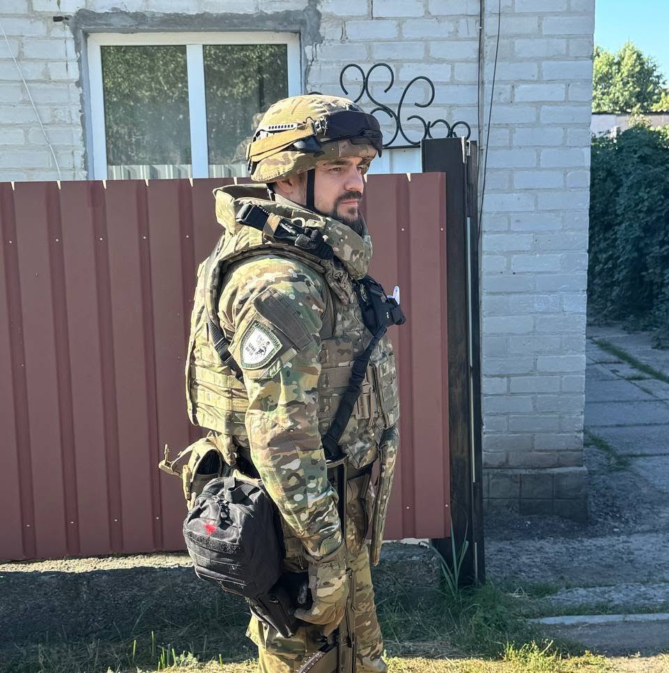 На фото чоловік у повному спорядженні стоїть біля будинку та паркану. Він одягнений у шолом, бронежилет і камуфляж, а на плечі видно нашивку підрозділу.