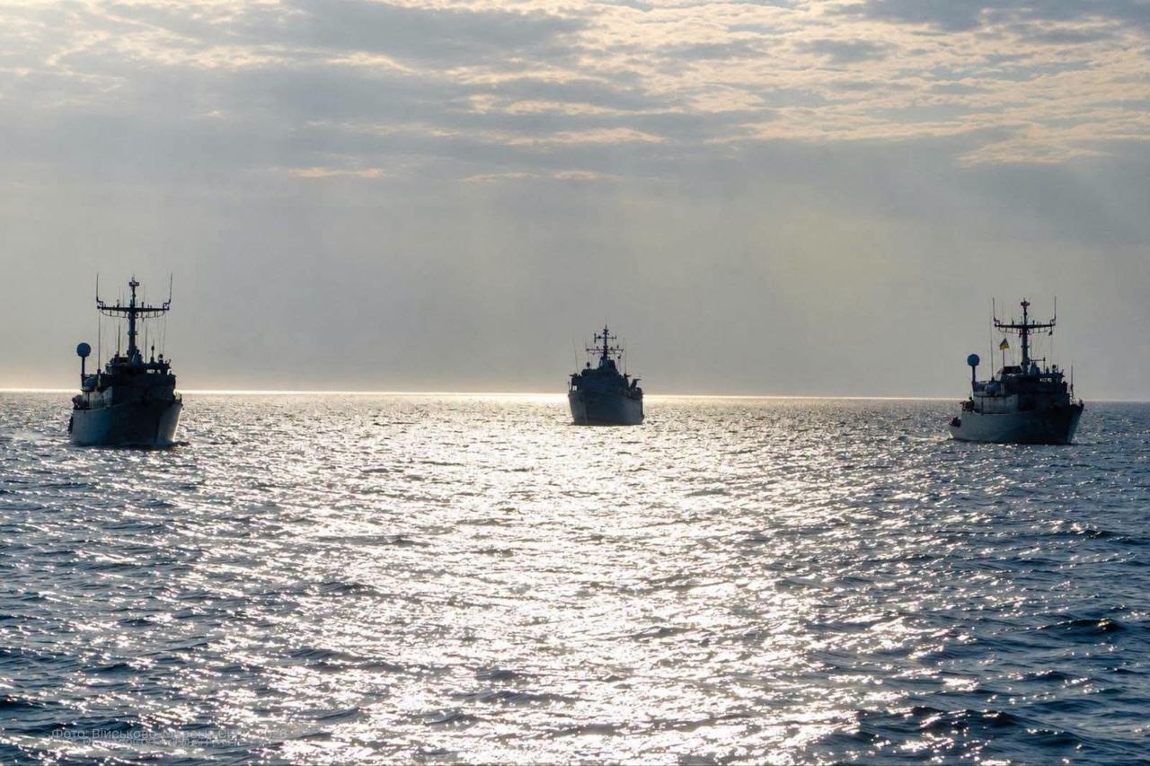 Три військові кораблі йдуть морем на значній відстані один від одного. Їхні темні силуети виділяються на тлі освітленої сонцем води та хмарного неба.