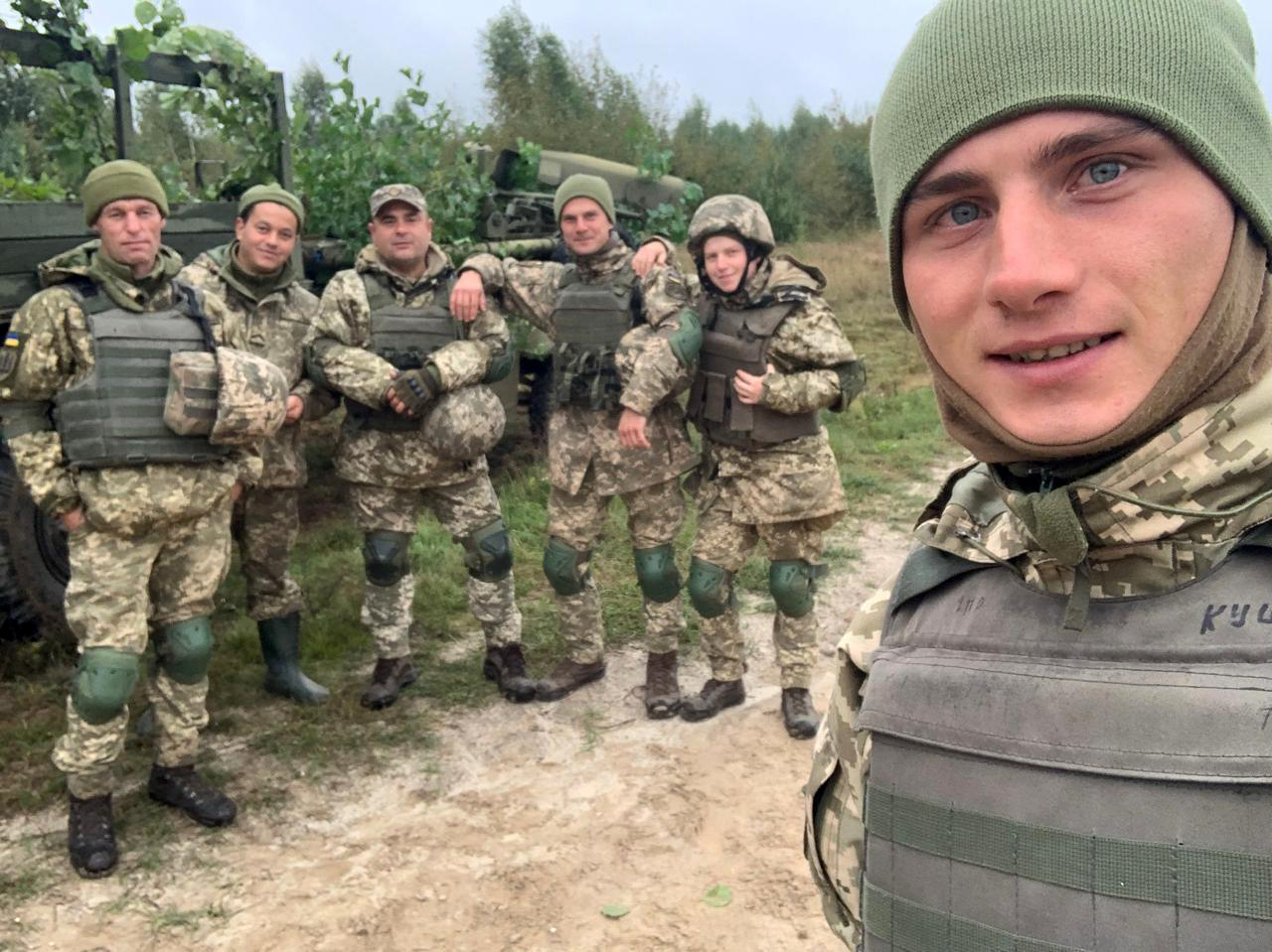 Група військовослужбовців у камуфляжі позує біля вантажівки та військової техніки на польовій дорозі. Один із них тримає камеру для селфі, а решта стоять у невимушеній атмосфері.