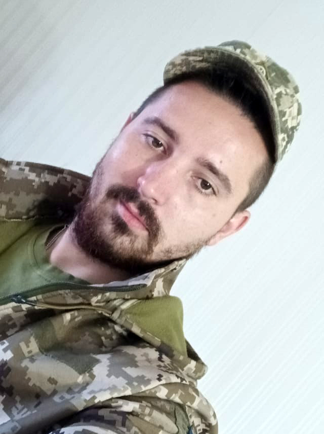 Чоловік у військовій формі зробив селфі у приміщенні. На ньому камуфляжна куртка й кепка, а вираз обличчя спокійний та зосереджений.