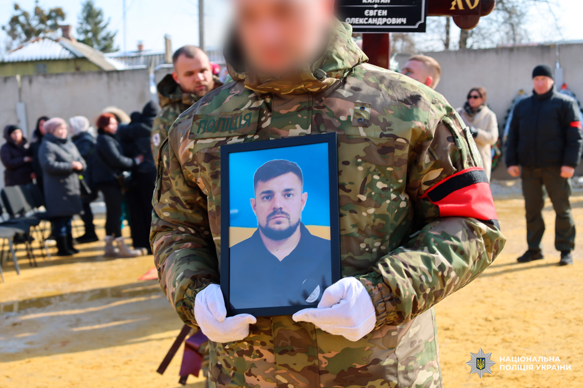 На фото людина у формі тримає портрет загиблого під час церемонії прощання або вшанування пам’яті. На задньому плані видно присутніх, а внизу кадру розміщений водяний знак Національної поліції України.