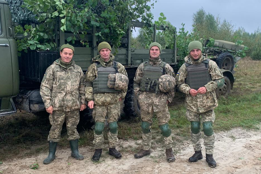 Четверо військових у камуфляжній формі стоять біля замаскованої вантажівки на узліссі. Поруч видно польову дорогу, траву та зелені гілки, якими прикрита техніка.