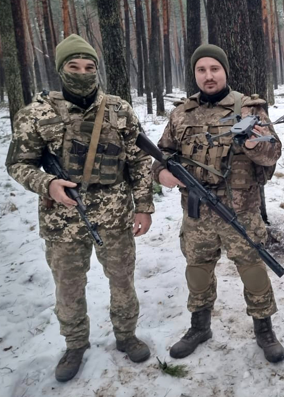 Двоє військовослужбовців стоять у засніженому лісі зі зброєю та безпілотником. Один із них тримає дрон у руці, що підкреслює роботу з безпілотними системами.