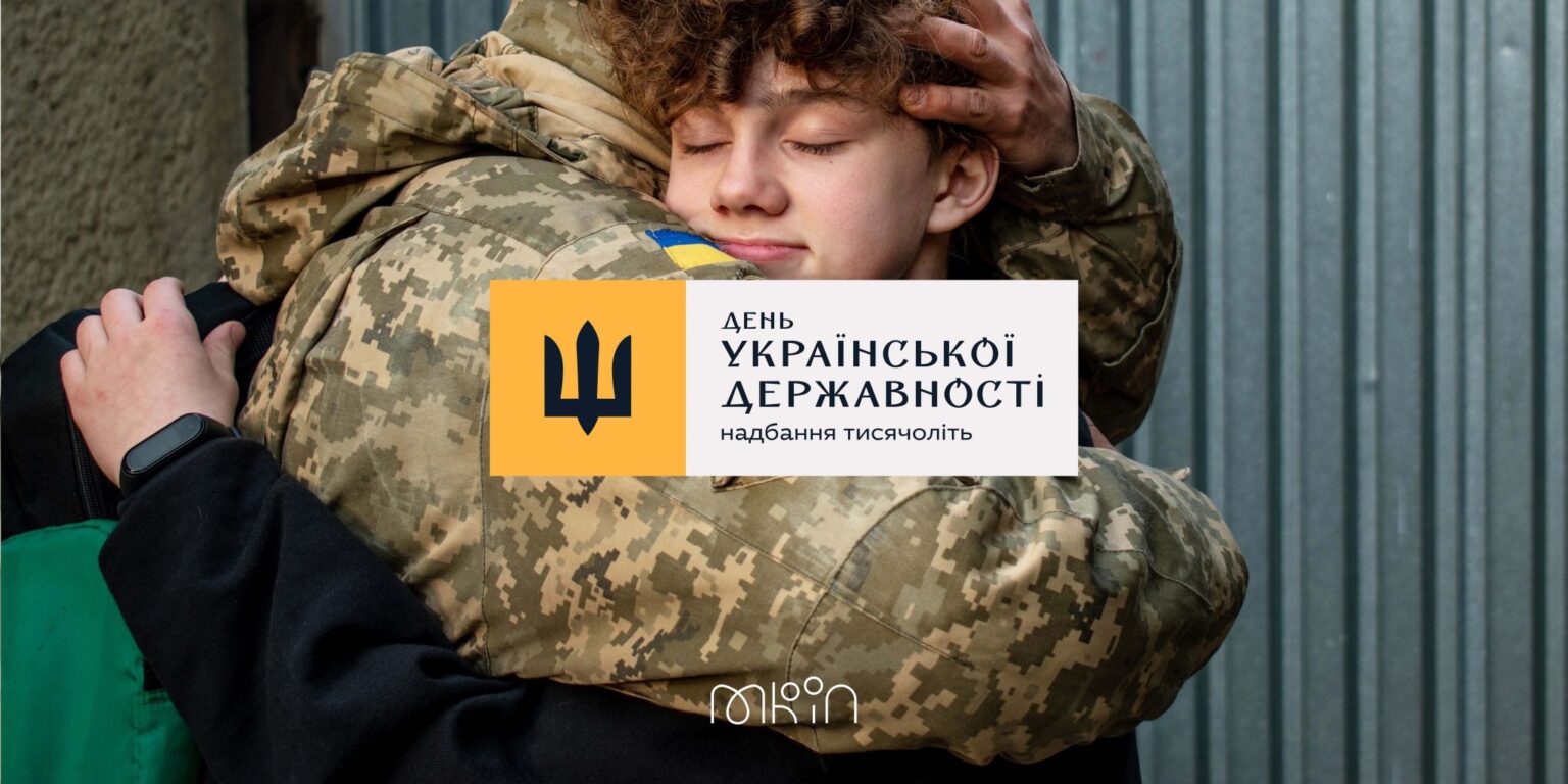 Український військовослужбовець у формі обіймає юнака, який із заплющеними очима притулився до його плеча. Поверх фотографії розміщено емблему Дня Української Державності та гасло «Надбання тисячоліть».