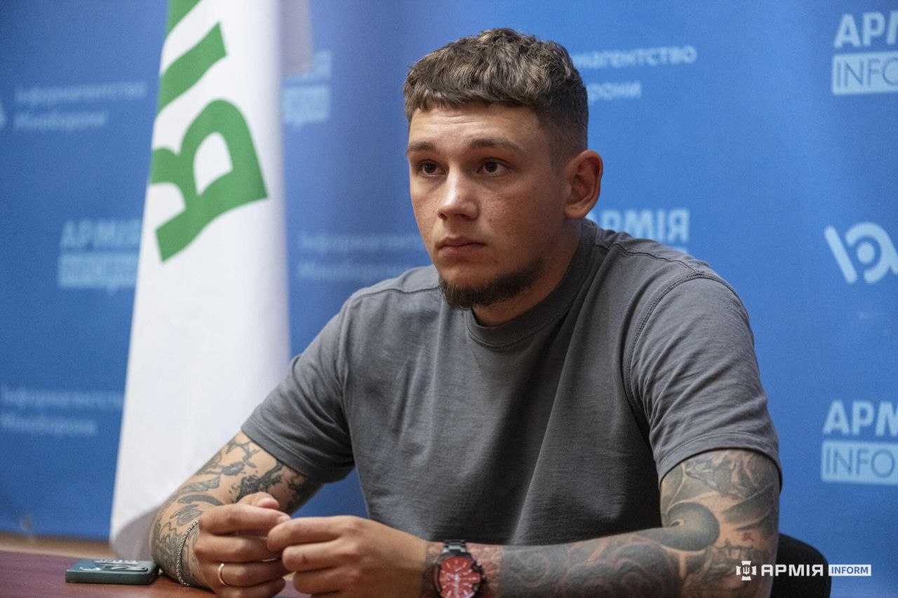Чоловік сидить за столом, тримаючи руки перед собою, та зосереджено дивиться на співрозмовника. Поруч лежить мобільний телефон, а позаду видно синій брендований банер і білий прапор.