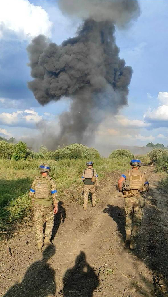 Троє військових ідуть польовою дорогою в напрямку великого стовпа диму після вибуху. У кадрі відчувається напружена бойова обстановка, а масштаби диму підкреслюють силу детонації.