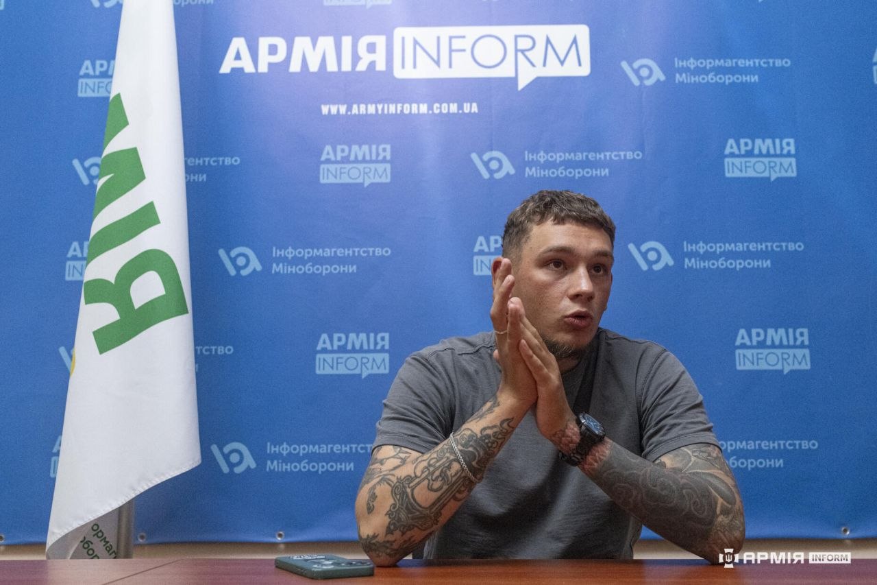 Чоловік сидить за столом і жестикулює складеними долонями під час розмови. Позаду нього розміщений великий синій банер із логотипами АрміяInform та інформаційного агентства Міністерства оборони.