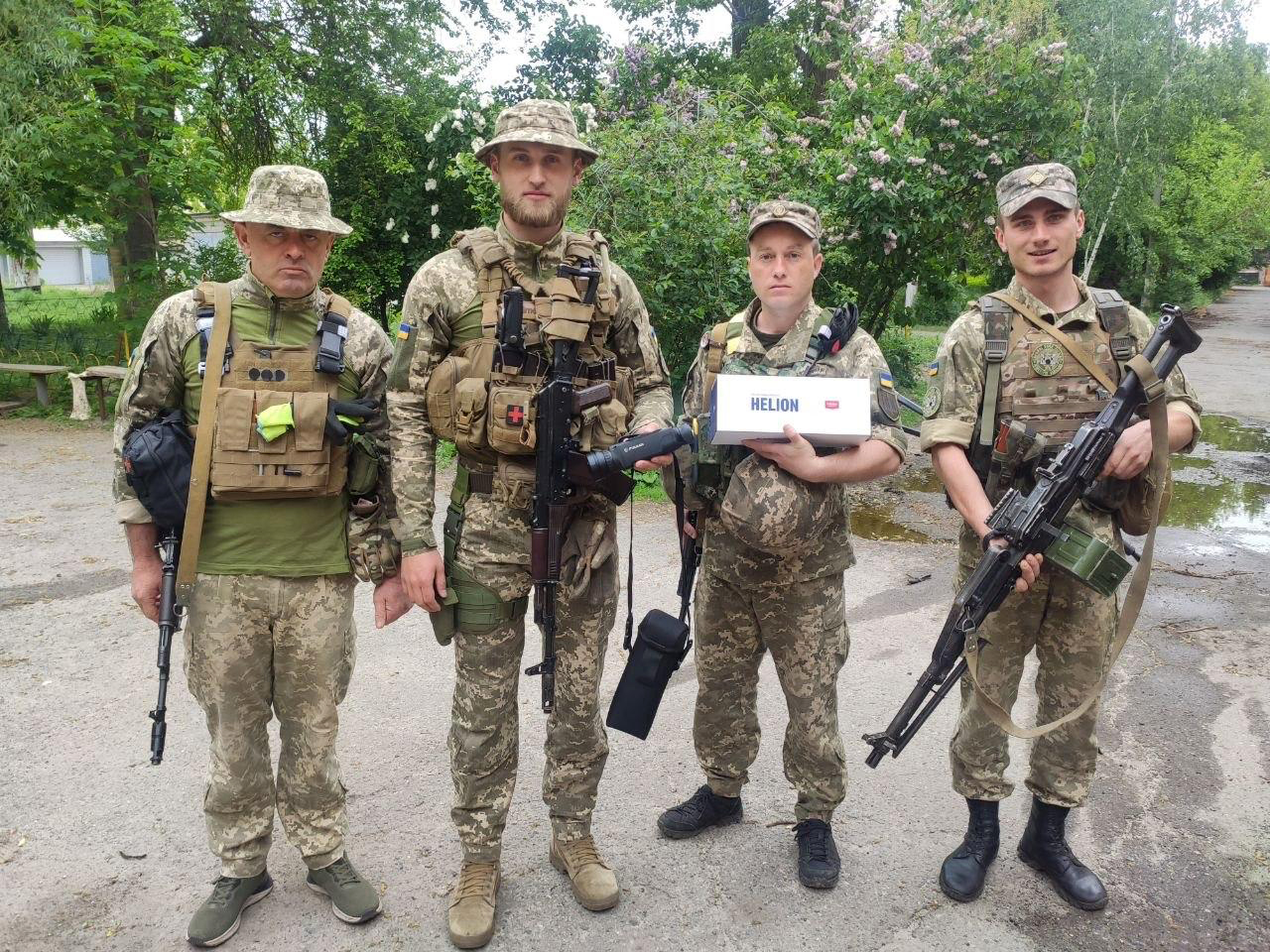 Четверо військових стоять на дорозі серед зелені, тримаючи зброю та коробку з написом HELION. Кадр виглядає як фіксація передачі або отримання обладнання для підрозділу.