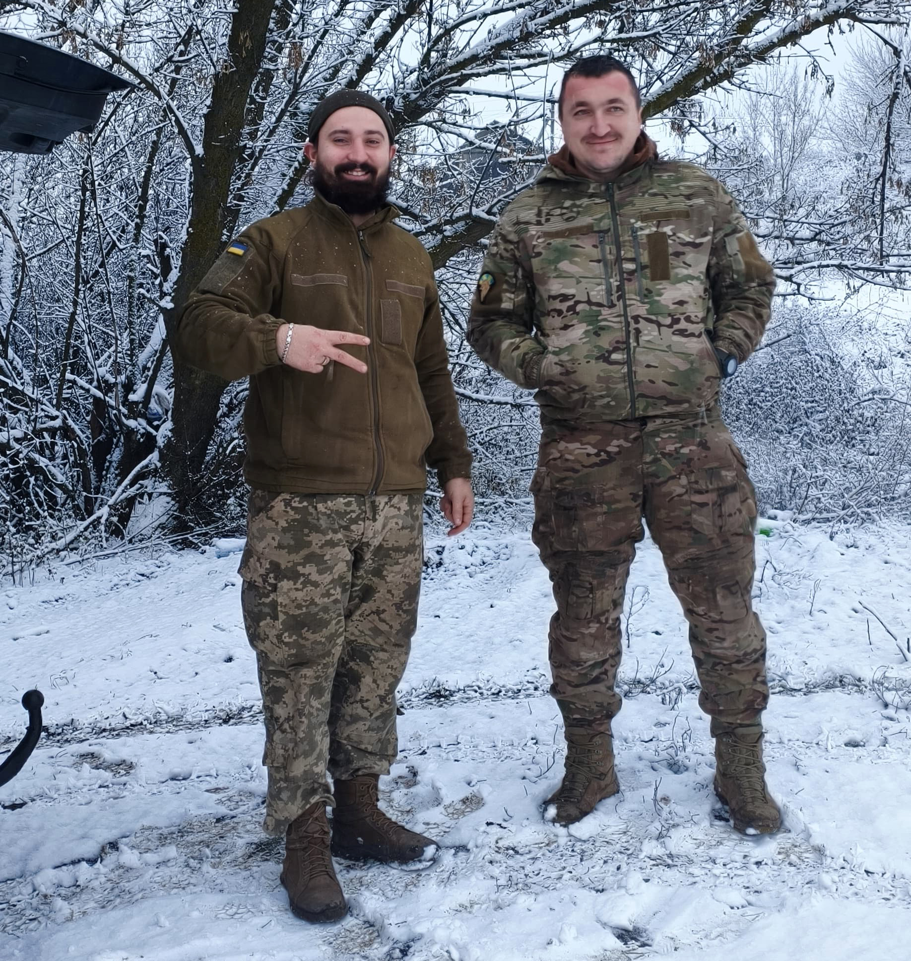 Двоє військових стоять на засніженій місцевості біля дерев. Вони усміхаються, один показує жест рукою, а зимовий фон додає кадру спокійного фронтового настрою.
