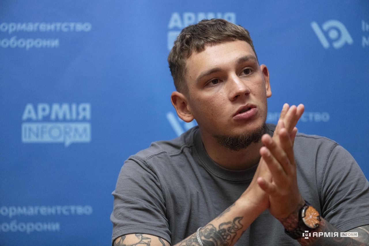 Чоловік говорить і супроводжує свої слова жестом складених долонь. Він дивиться вбік на співрозмовника, сидячи перед синім інформаційним банером.