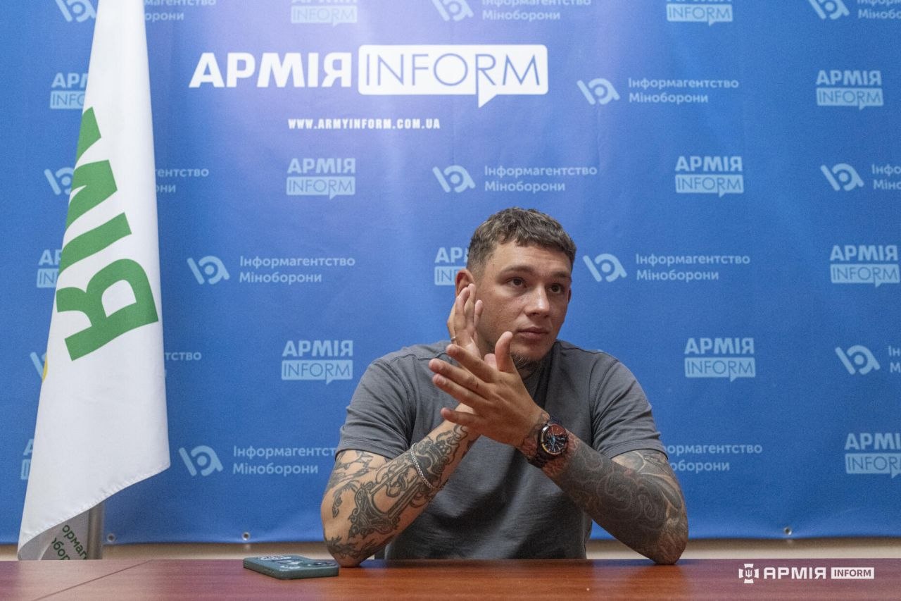 Чоловік сидить за столом і жестикулює руками під час розмови. Позаду розміщений синій брендований банер АрміяInform, ліворуч стоїть прапор, а на столі лежить смартфон.