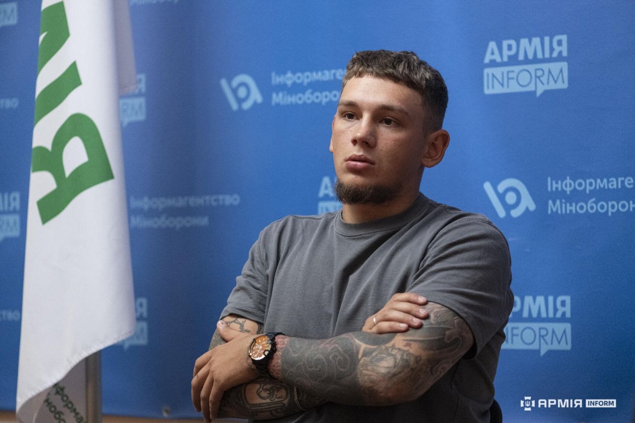 Чоловік зі схрещеними руками уважно слухає співрозмовника. Його татуйовані передпліччя та годинник добре помітні на тлі синього банера АрміяInform.