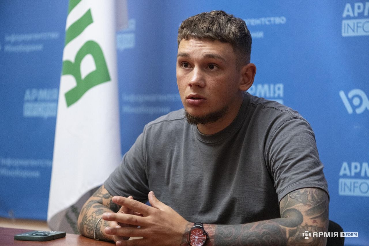Чоловік сидить за столом зі складеними руками та говорить, дивлячись у бік співрозмовника. Позаду видно прапор і синій брендований банер АрміяInform, а на столі лежить смартфон.