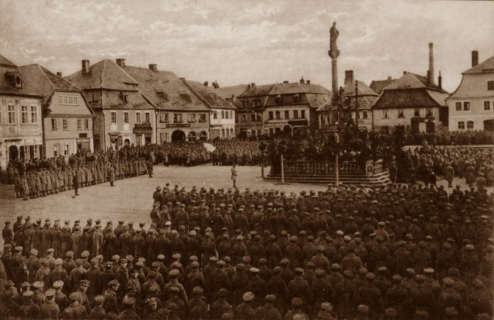 Історична світлина показує масове зібрання військових і цивільних на центральній площі європейського міста. Люди стоять навколо пам’ятної колони, а вздовж країв площі вишикувані військові підрозділи.