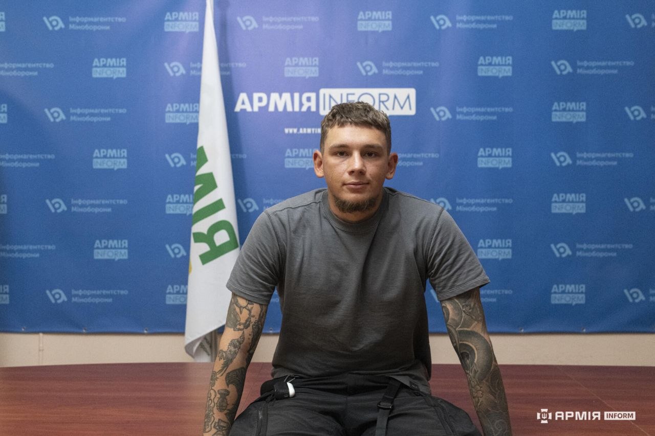 Чоловік сидить у центрі за столом, спираючись руками на його край. Він дивиться прямо перед собою, а позаду видно прапор і брендовану стіну АрміяInform.