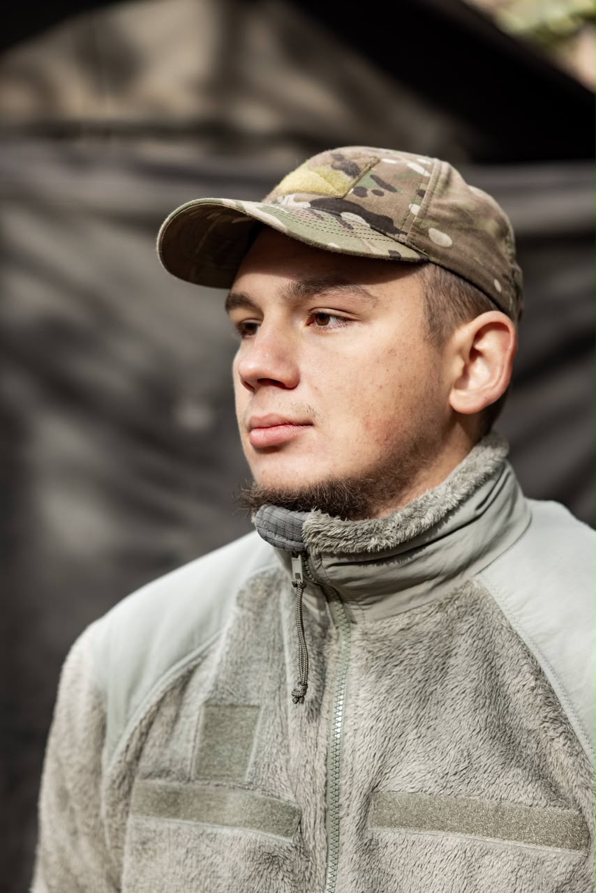 Чоловік у камуфляжній кепці та теплому флісовому одязі дивиться вбік із серйозним виразом обличчя. Розмитий темний фон акцентує увагу на його портреті.