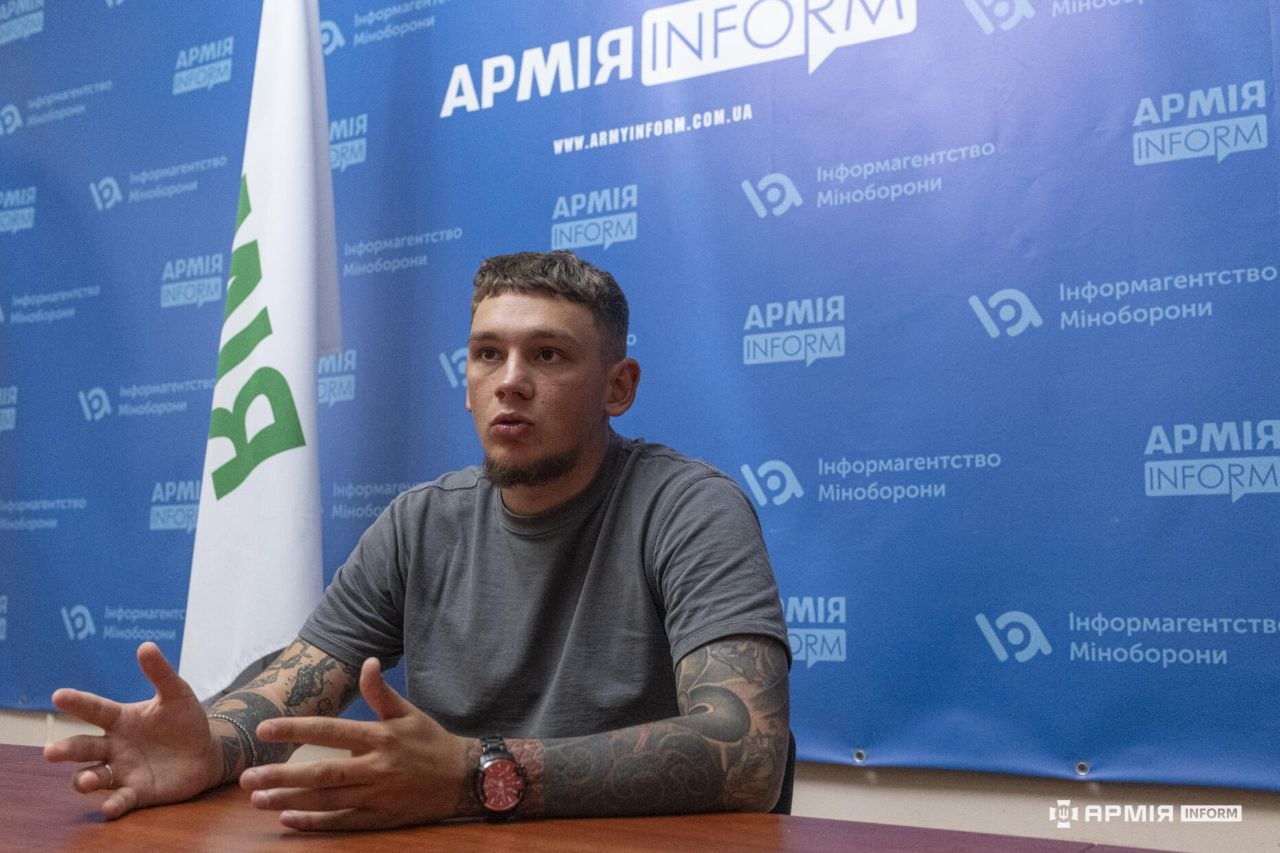 Чоловік сидить за столом і широко жестикулює руками, пояснюючи щось під час розмови. Позаду розташовані прапор і синій банер з логотипами інформаційного агентства АрміяInform.
