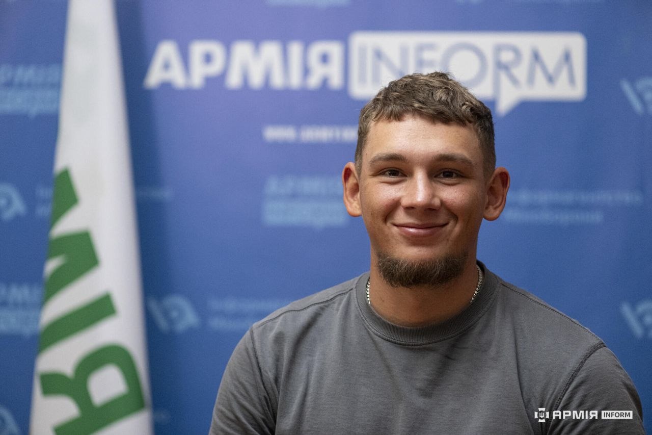 Чоловік усміхається, дивлячись прямо перед собою. Портрет знятий крупним планом на тлі прапора та розмитого брендованого банера АрміяInform.