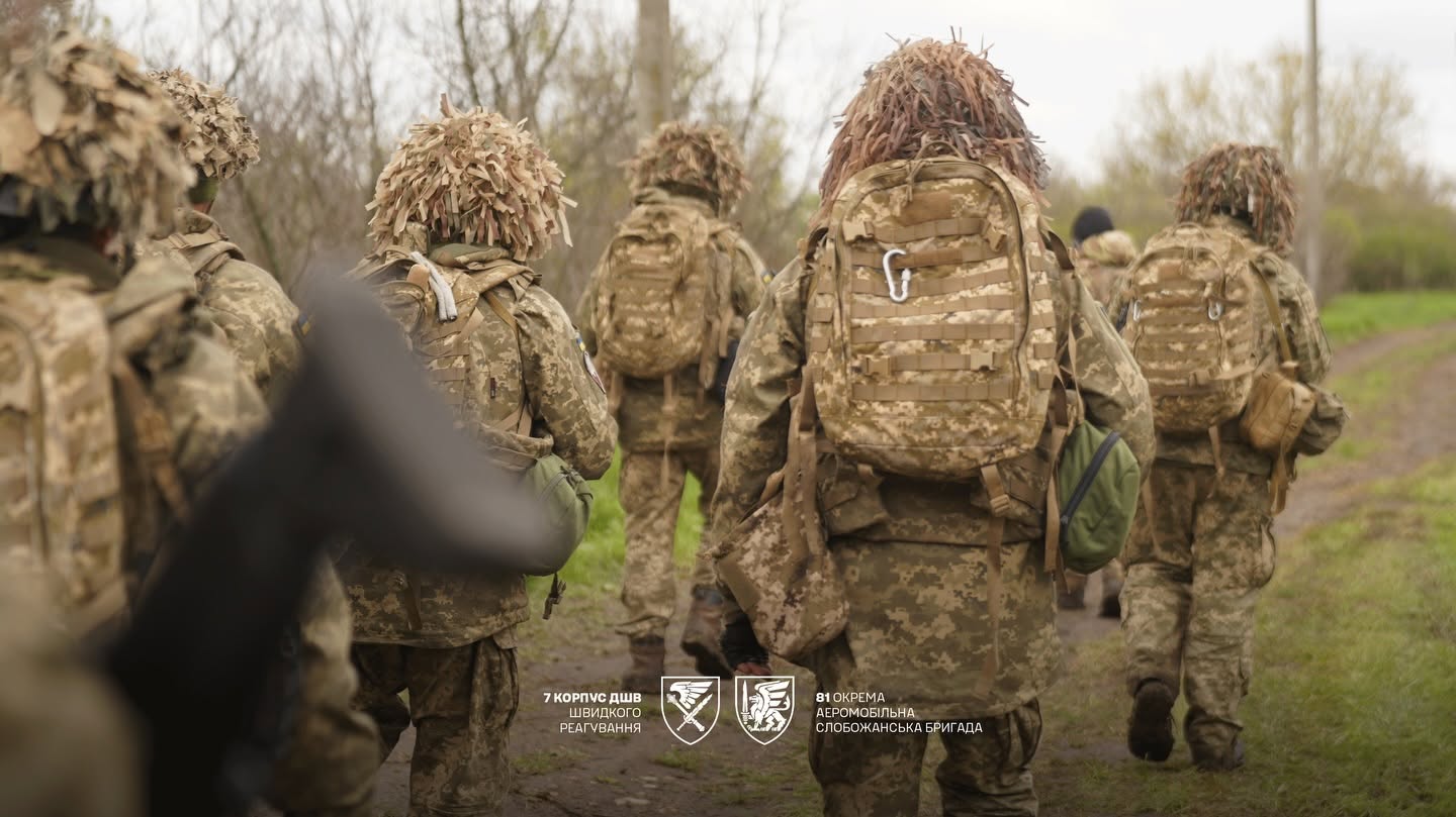 Група військовослужбовців у камуфляжі та маскувальних головних уборах рухається ґрунтовою дорогою, несучи наплічники й особисте спорядження. Унизу кадру розміщені емблеми 7-го корпусу швидкого реагування ДШВ та 81-ї окремої аеромобільної Слобожанської бригади.