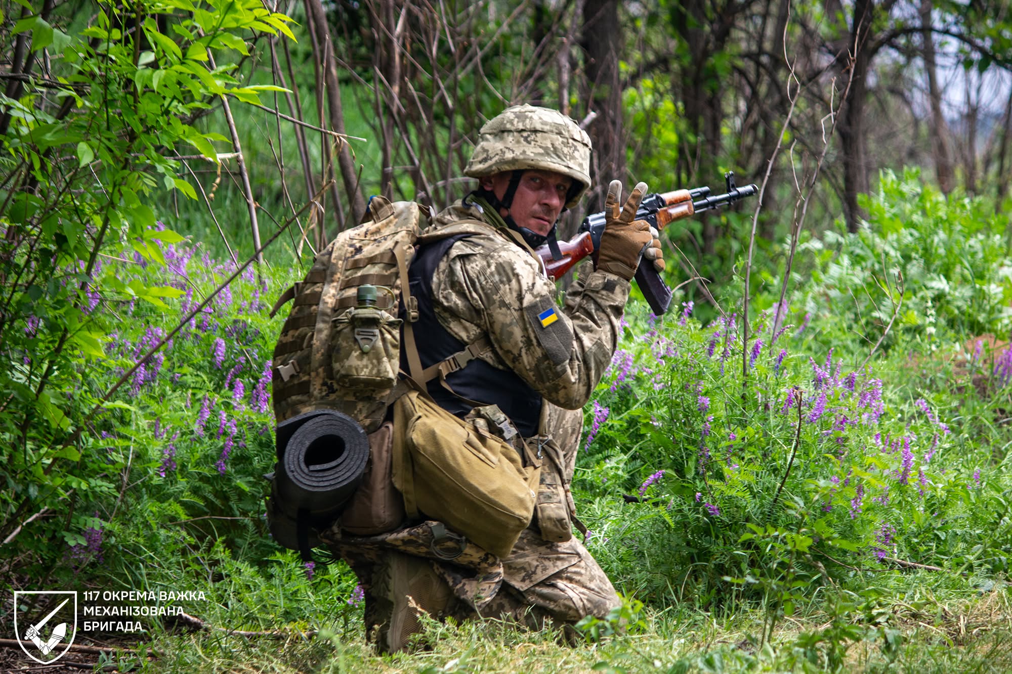 Український військовослужбовець у камуфляжі та шоломі стоїть на коліні серед густої зеленої рослинності, тримаючи в руках автомат. На його формі видно синьо-жовтий прапор, а внизу зображення розміщений підпис 117-ї окремої важкої механізованої бригади.