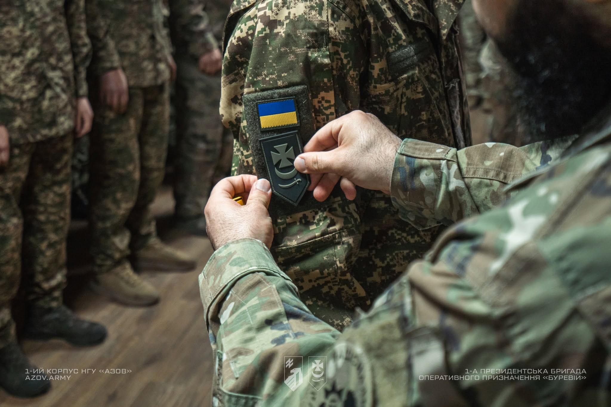 Військовому прикріплюють нарукавний знак підрозділу під час шикування, поруч видно прапор України на формі. Кадр передає момент офіційного вручення або посвяти, з акцентом на символіці служби та належності до бригади.